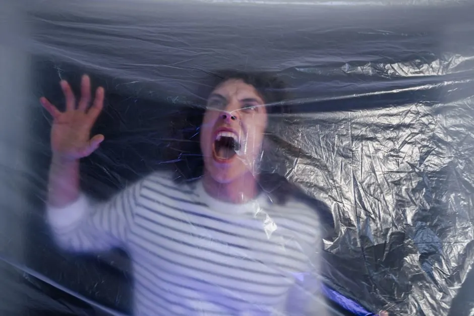 Man in striped shirt screaming behind translucent plastic foil, conveying fear.